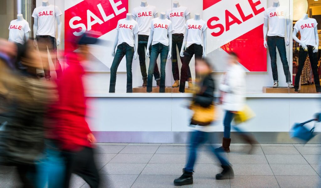 A shop window full of sales signs with shoppers walking past on a busy street