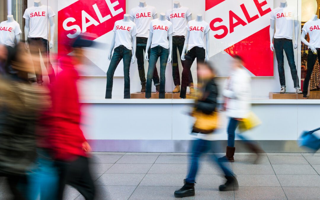 A shop window full of sales signs with shoppers walking past on a busy street